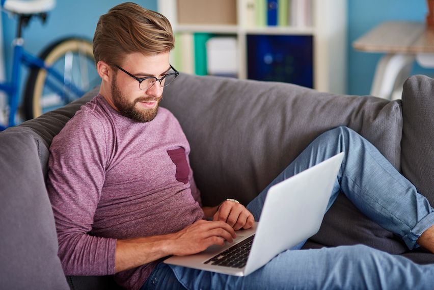 young man with glasses working from home, sitting on his couch, looking at his notebook.