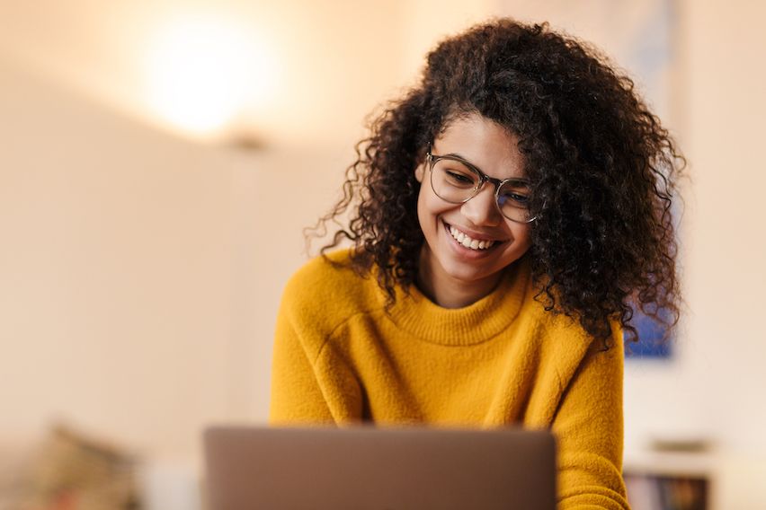Smiling woman with black curly hair in a warm yellow pullover, crafting personalized email content on her laptop.