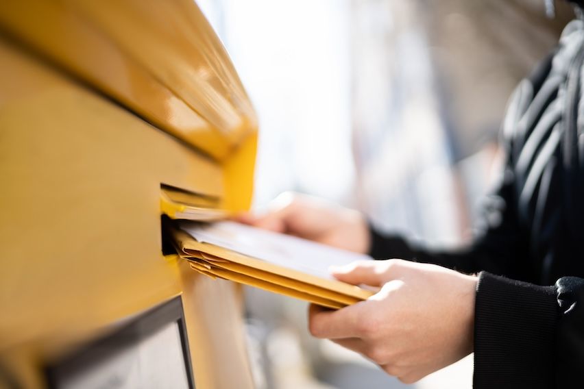 Hands inserting envelopes into a yellow letterbox, symbolizing strategic timing in targeted mail marketing campaigns.