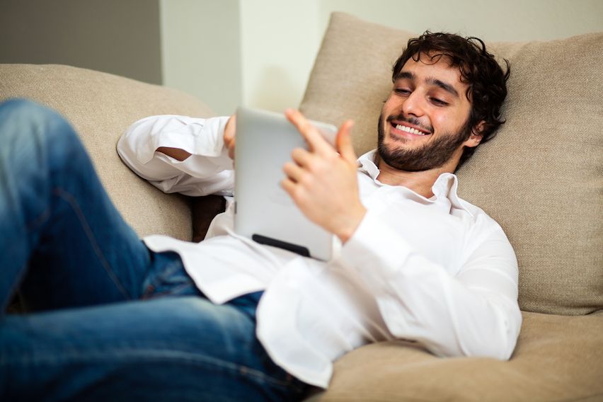 Happy young man lounging on a couch, effortlessly setting up an email marketing campaign on his tablet.